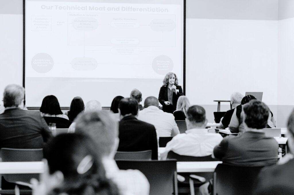 A woman stands in front of a presentation screen, addressing a seated audience in a conference room. The slide behind her displays a diagram related to technical analysis and differentiation.